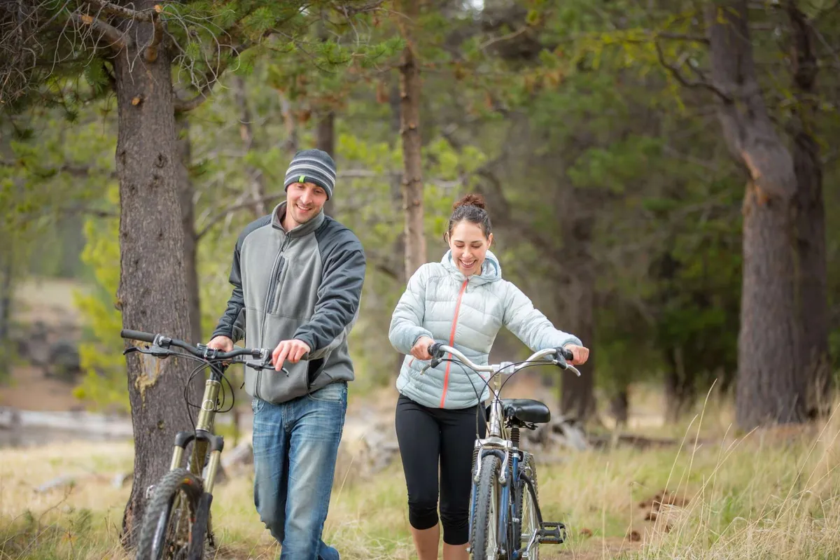 An exterior shot of a couple walking with bicycles along a tree-lined path with open grassy areas, conveying an active, social lifestyle. The natural setting suggests easy access to local trails and the outdoors, mirroring the adventure and exploration described in the section.