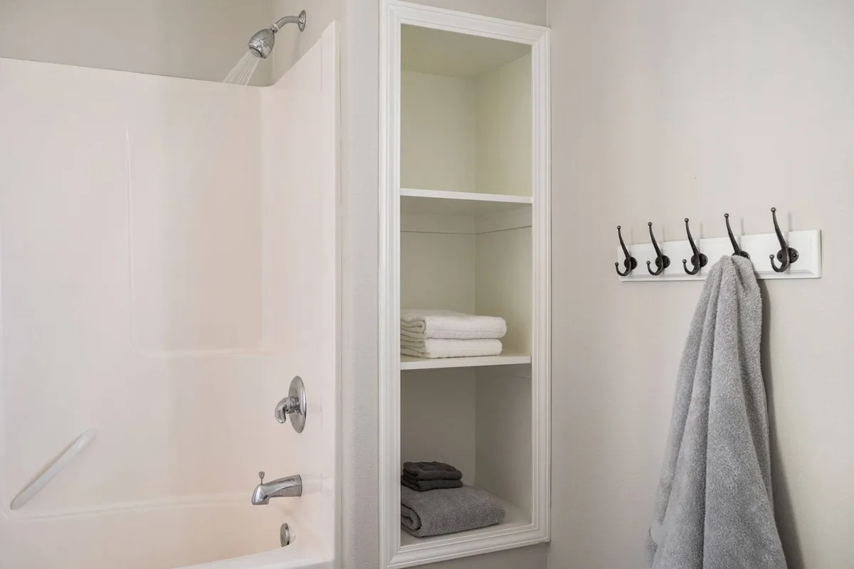 A soothing bathroom setting with a spacious bathtub, plush towels, open shelves for storage, and convenient wall hooks. The ambience is calm and spa-like, emphasizing self-care and relaxation.
