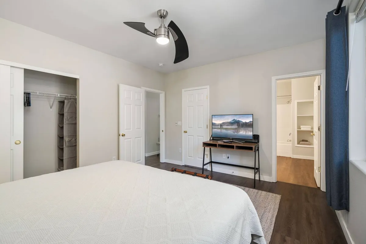 A tranquil bedroom scene centered on a comfortable bed facing a television, with a ceiling fan overhead. The arrangement suggests both relaxation and entertainment, and the ceiling fan is prominently featured, aligning with amenities mentioned in the content.