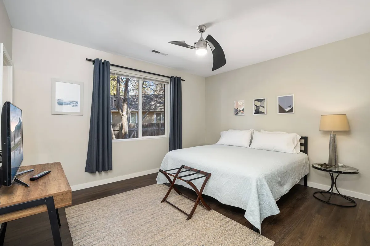 A peaceful bedroom featuring a cozy bed with crisp linens, a large window letting in soft natural light, a nightstand with a lamp, subtle artwork on the wall, and a plush rug underfoot. The scene evokes a sense of relaxation and quiet retreat, perfect for unwinding after a day outdoors.