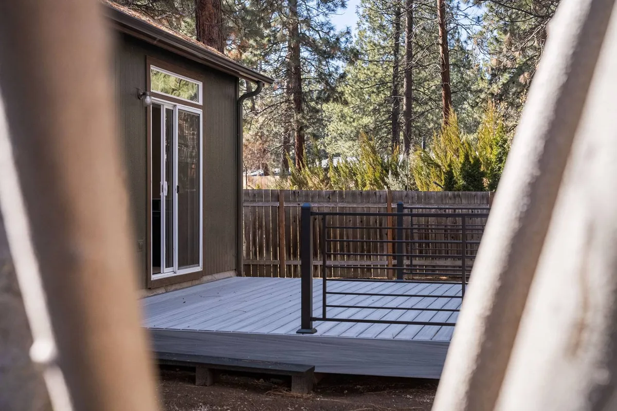 A view of the deck adjacent to the house, framed by windows and lush trees. The deck is spacious, ideal for outdoor relaxation or gathering, and the surrounding greenery hints at privacy and a connection to nature.