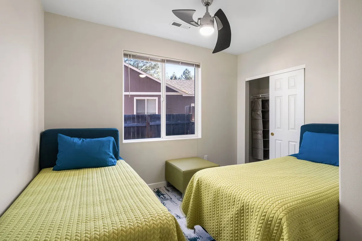 A bright and airy guest bedroom with two twin beds, each neatly made, positioned beneath a large window with views outside. A ceiling fan is visible overhead, and the room feels inviting—ideal for families or friends sharing the space.