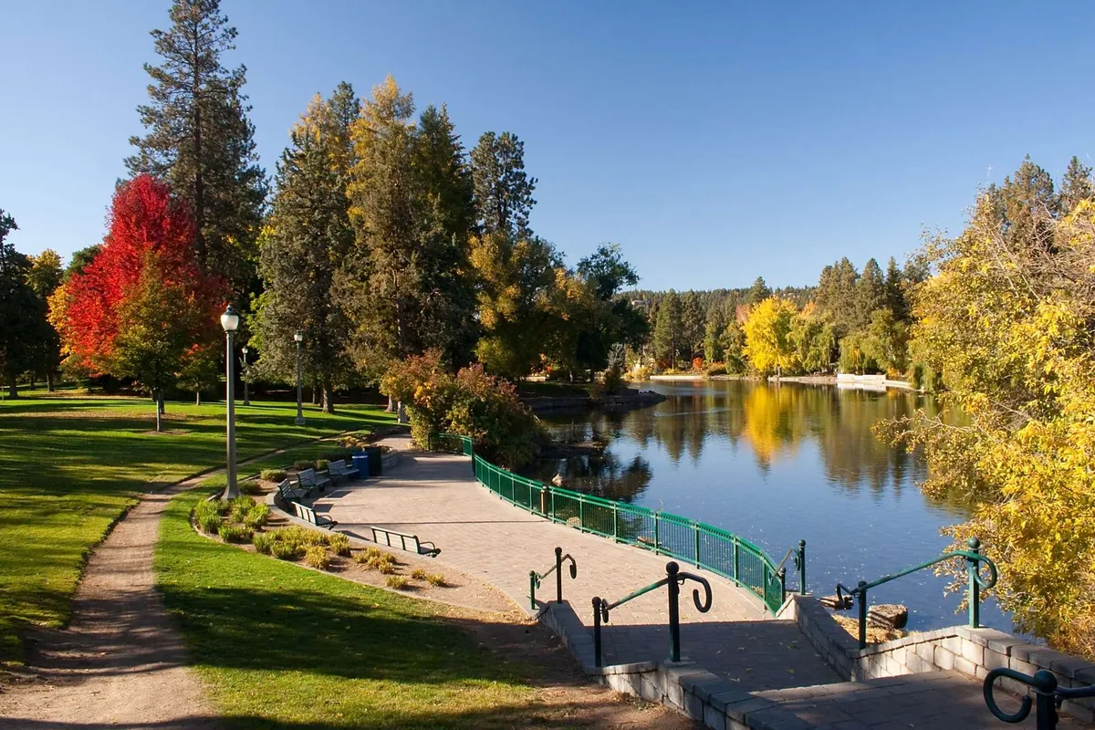 A tranquil lake scene bordered by colorful trees and walking paths, featuring park benches—an inviting spot for picnics, birdwatching, or peaceful walks after sampling the town’s offerings. The calm, scenic setting ties into Bend’s reputation for natural beauty.