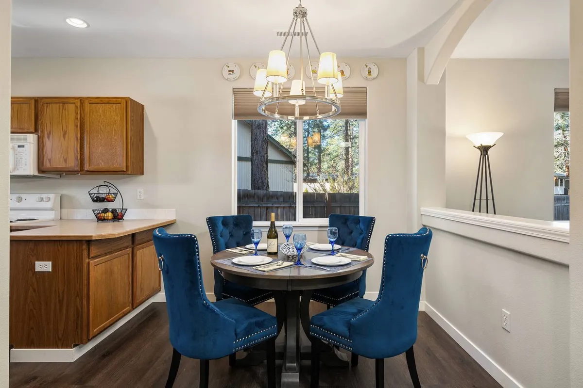 An elegant dining area with a round table, blue velvet chairs, a stylish chandelier, and a window allowing natural light to stream in. The setup is intimate yet welcoming, ideal for family dinners or sharing stories over meals.