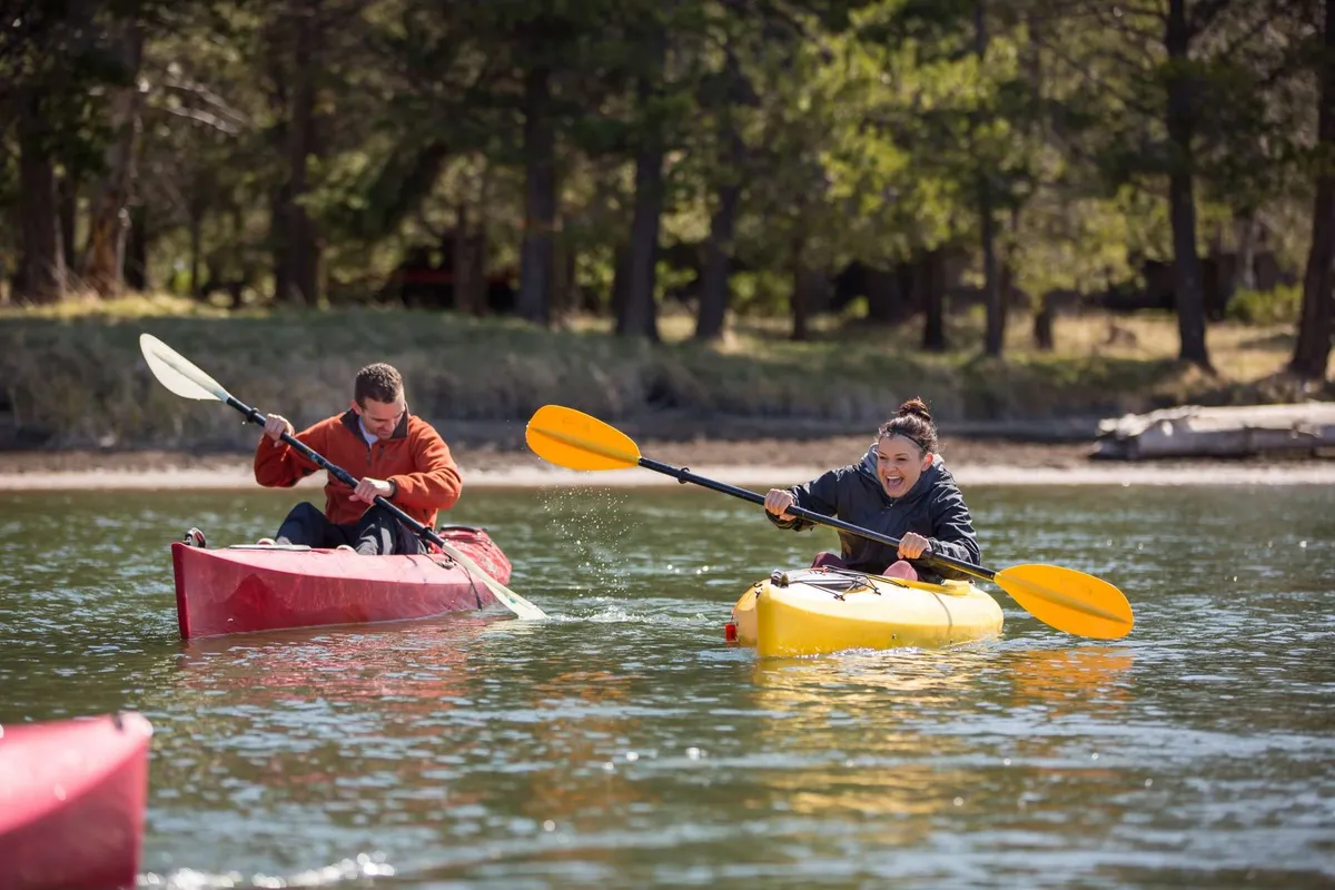 Two people kayak brightly colored boats across a reflective body of water, surrounded by lush greenery. The mood is energetic and joyful, evoking a sense of summer fun and water-based adventure.