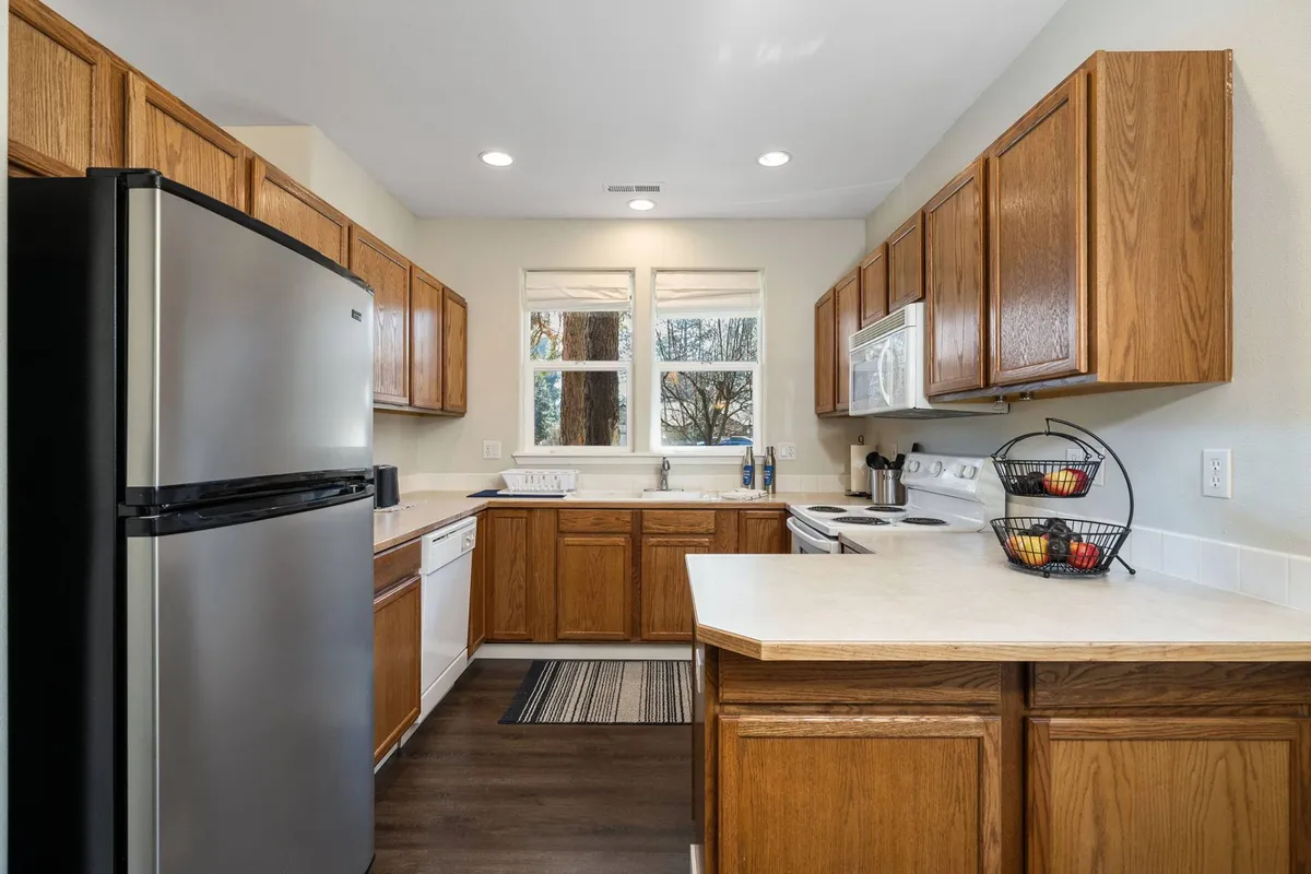 A modern kitchen with stainless steel appliances, wooden cabinets, a central kitchen island, a countertop adorned with a fruit basket, and a window providing outdoor views. The layout encourages socializing and group meal preparation.
