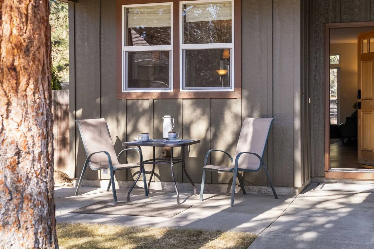 A small outdoor table is set with a coffee pot and cups, flanked by two chairs on a deck surrounded by greenery. The setting suggests a quiet morning or evening spent outdoors, with ample space and privacy provided by the surrounding trees.